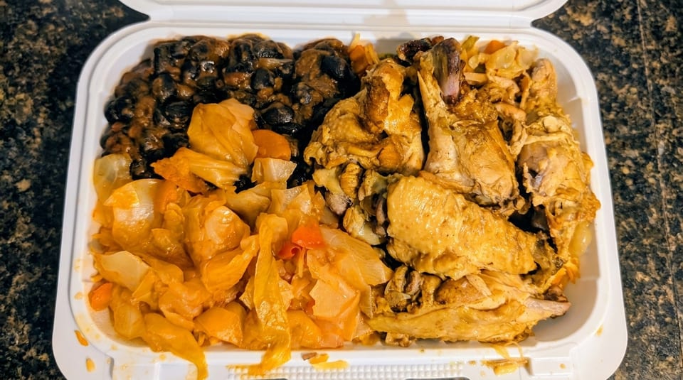 A takeout container filled with seasoned chicken, black beans, and stewed cabbage on a dark counter.